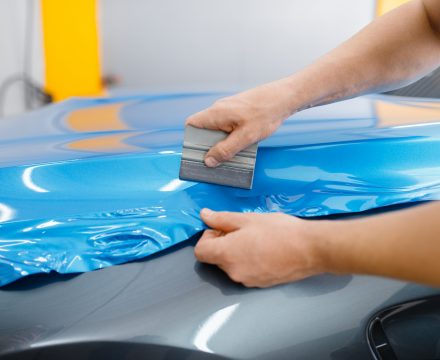 Car wrapping, mechanic with squeegee installs protective vinyl foil or film on vehicle hood. Worker makes auto detailing. Automobile paint protection coating, professional tuning
